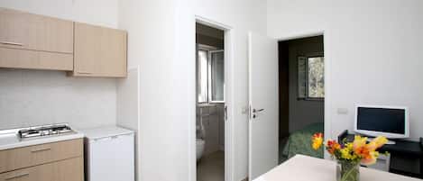 A compact kitchen with a refrigerator, stove, and cabinets, a small bathroom with a toilet and sink, and a desk with a computer and a vase of flowers.