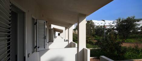 Un espacio al aire libre techado, con piso de baldosas, pared blanca y una puerta con contraventanas.