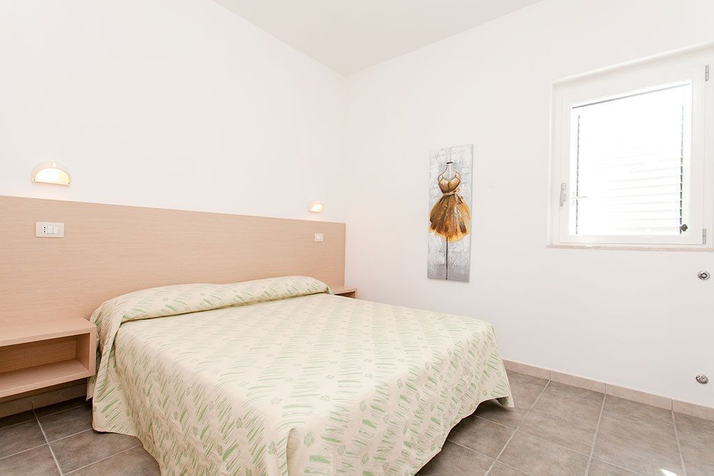 Un dormitorio con cama, cabecera, lámpara de pared, ventana y un cuadro.