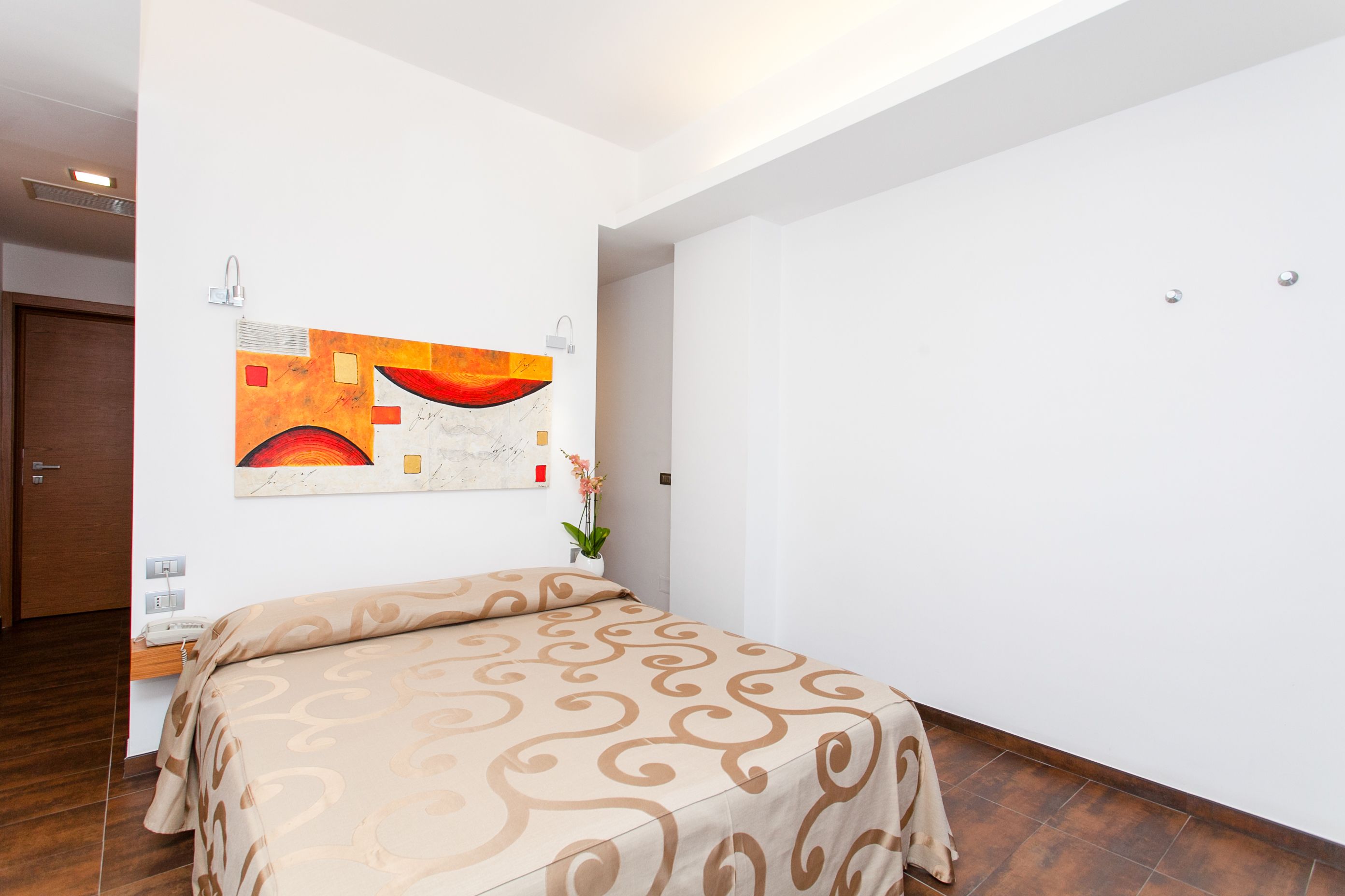 A hotel room with a bed, a nightstand, a wall-mounted artwork, and a potted plant.