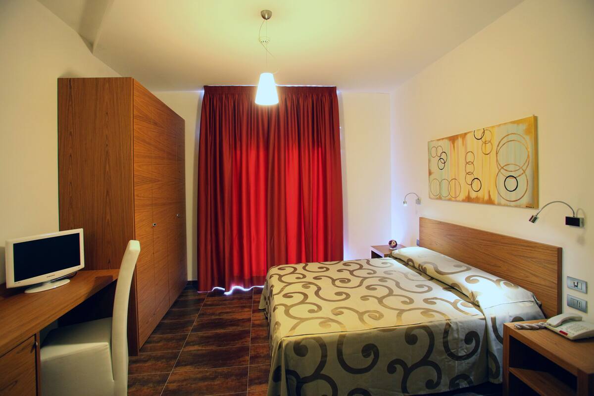 A hotel room with a bed, a desk with a computer, a chair, a TV, and a red curtain.