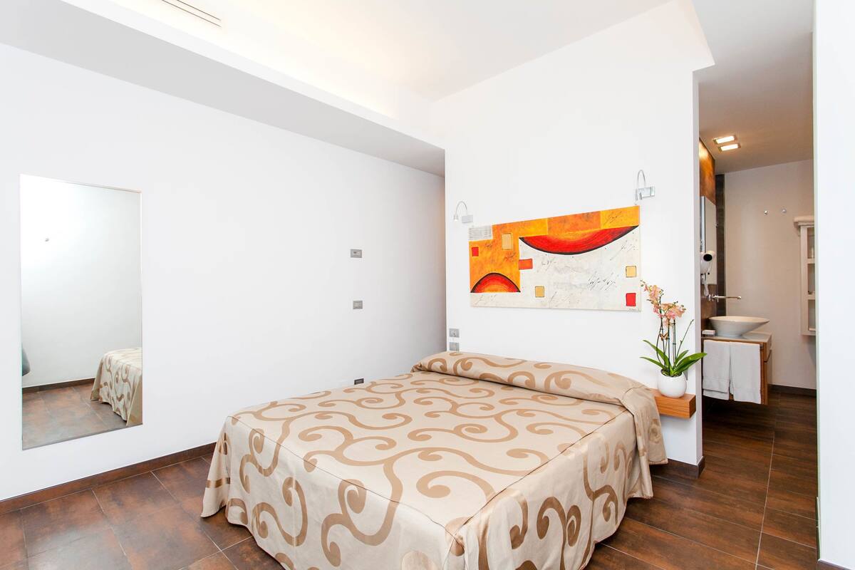 A bedroom with a bed, a mirror, a wall art piece, and a bathroom visible through an open door.