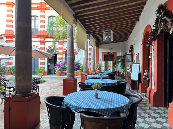 An outdoor patio with round tables and chairs, potted plants, and a view of a building with red and white walls. - Mesón de Santa Elena Hotel Galeria (Mascota)