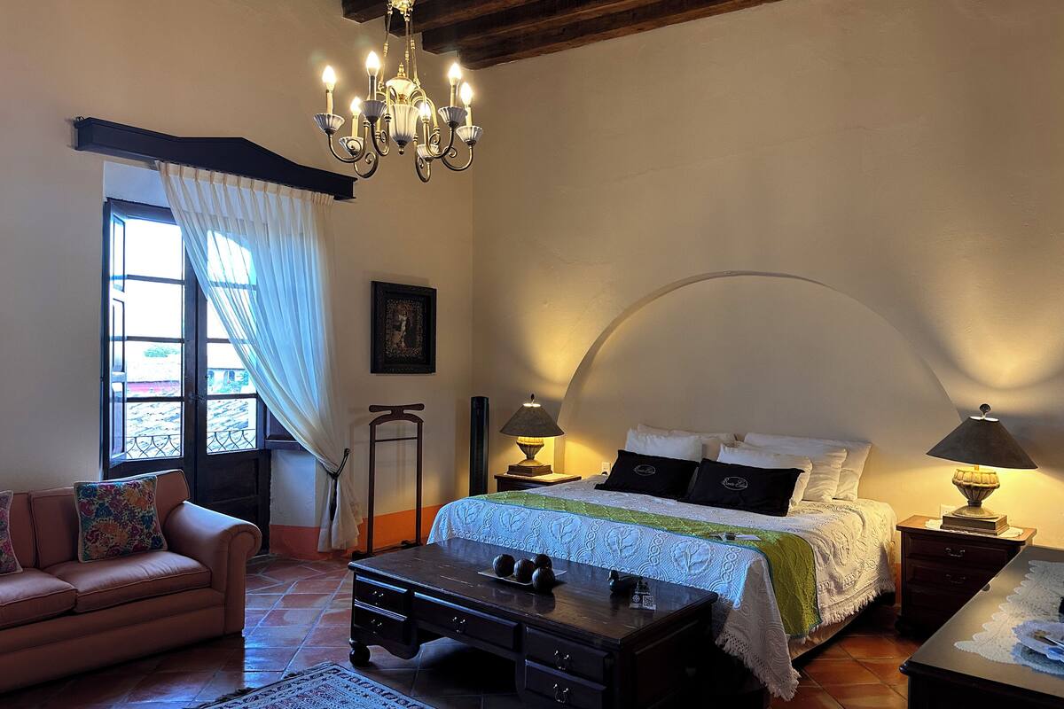 Une chambre à coucher comprenant un lit, des tables de chevet, un bureau, une chaise, un lustre et une fenêtre avec des rideaux.