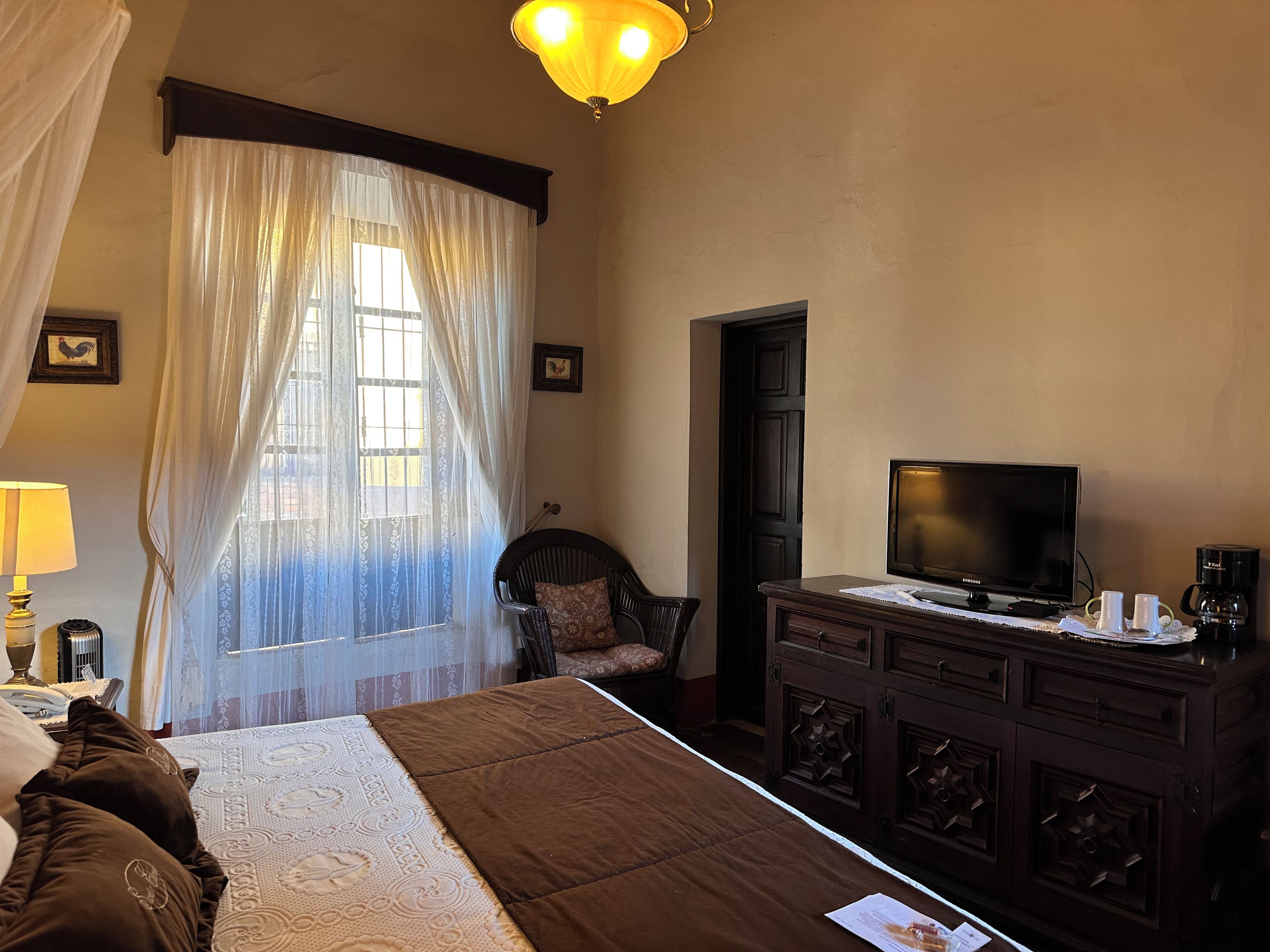 A hotel room with a bed, a television, a desk with a coffee maker, a chair, and a window with curtains.