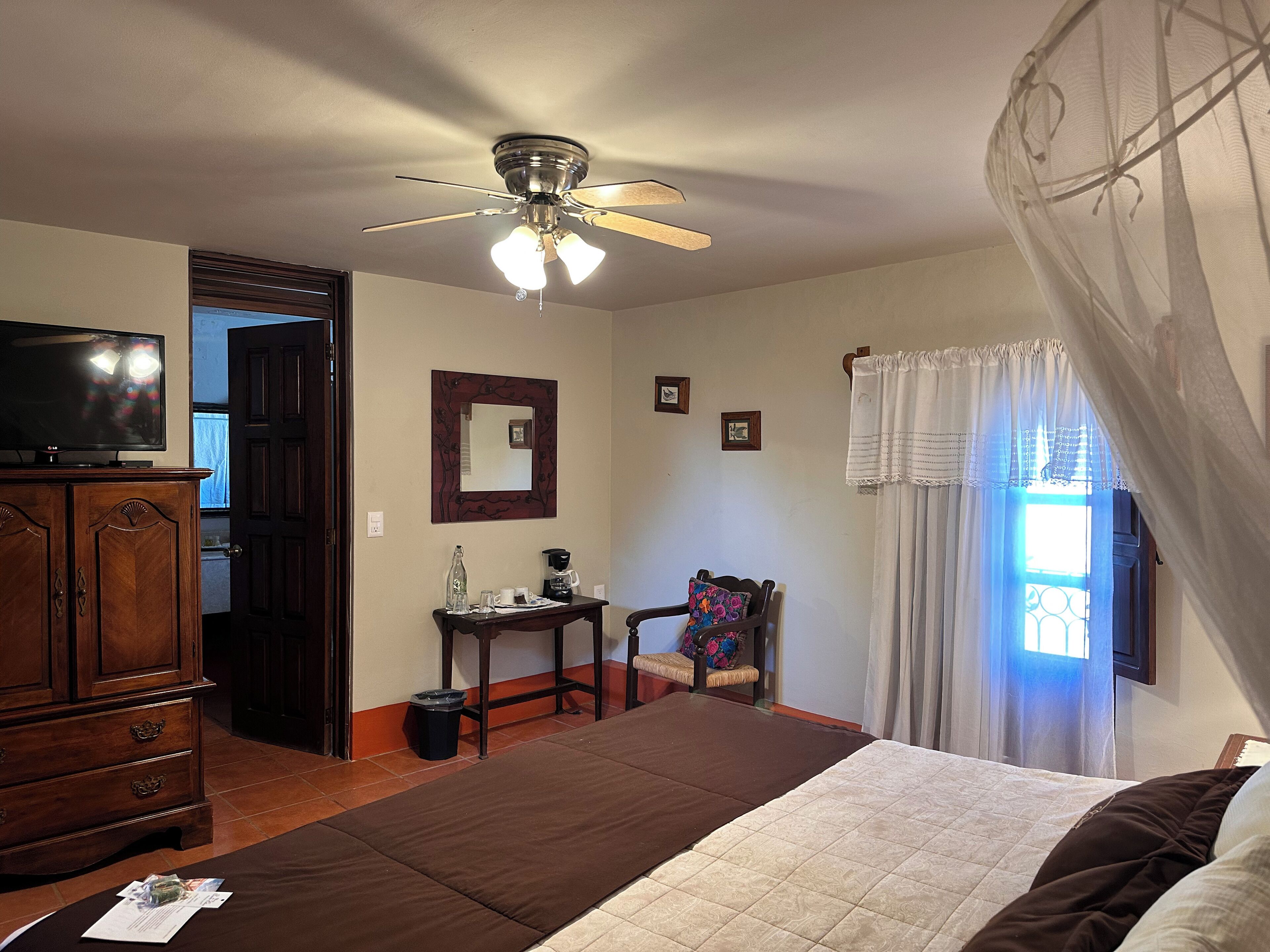 Un dormitorio con cama, ventilador de techo, televisor, armario de madera, una mesita con cafetera y ventana con cortinas.