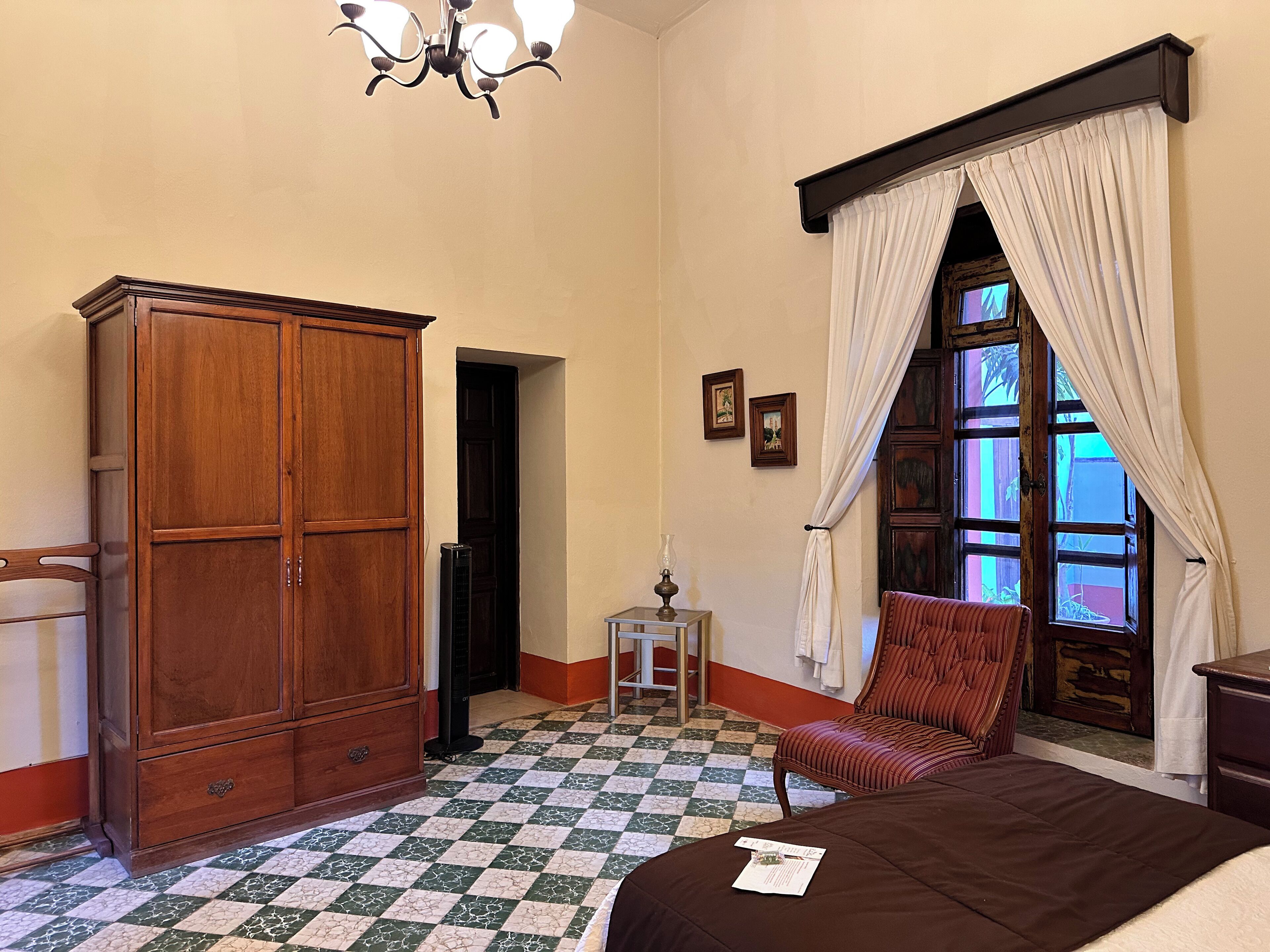 Habitació amb armari de fusta, llit amb manta marró, taula petita amb làmpada i finestra amb cortines.