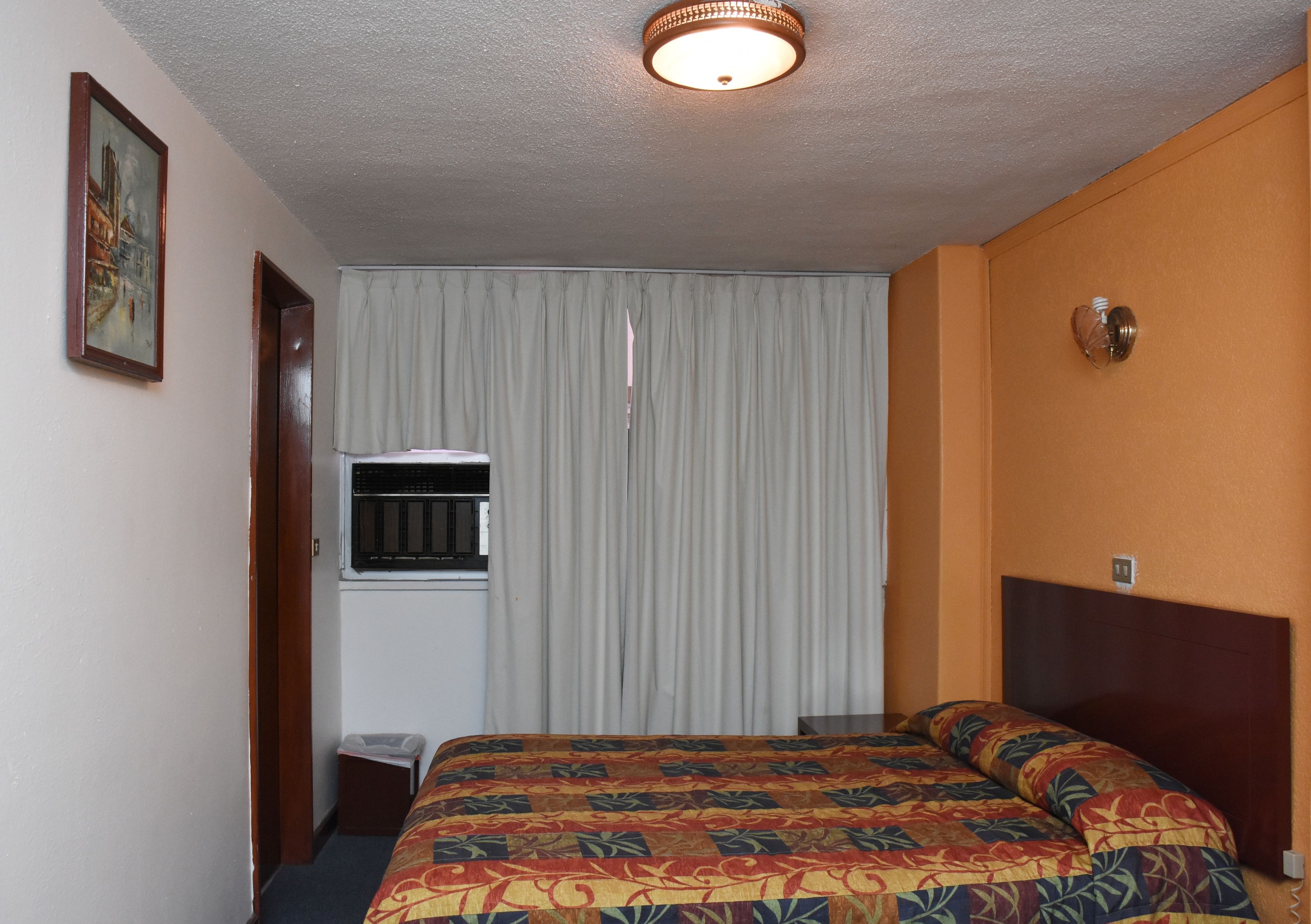 Habitación de hotel con cama, ventana con cortinas, puerta y lámpara fijada a la pared.