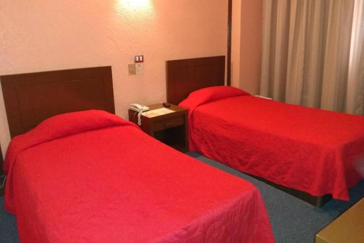 A hotel room with two single beds, each with a red bedspread, a wooden headboard, a bedside table with a lamp, and a curtain on the window.