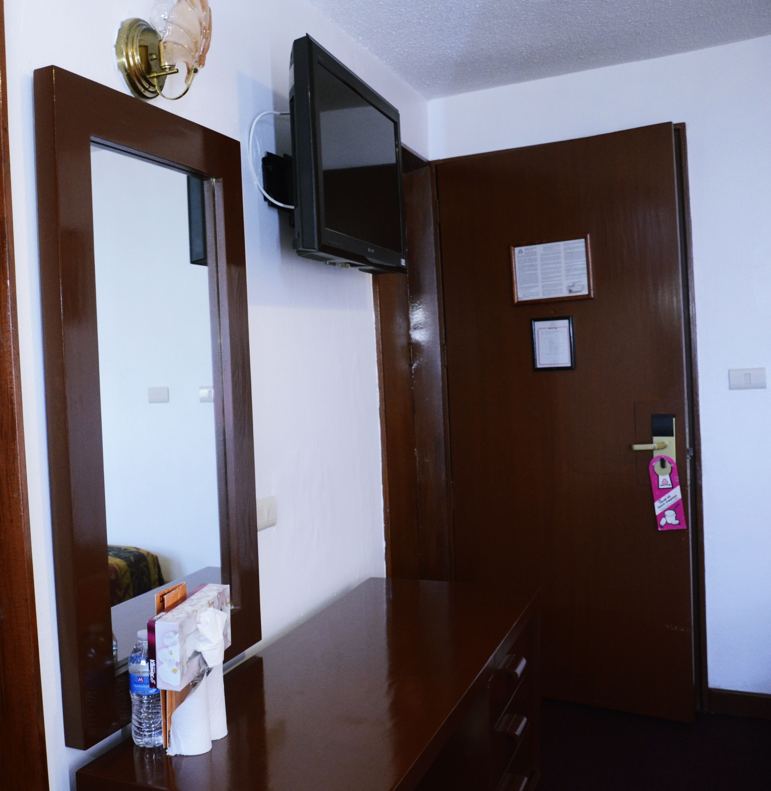 Une chambre d’hôtel avec une porte en bois, un téléviseur fixé au mur, un miroir et une petite table avec des articles de toilette.