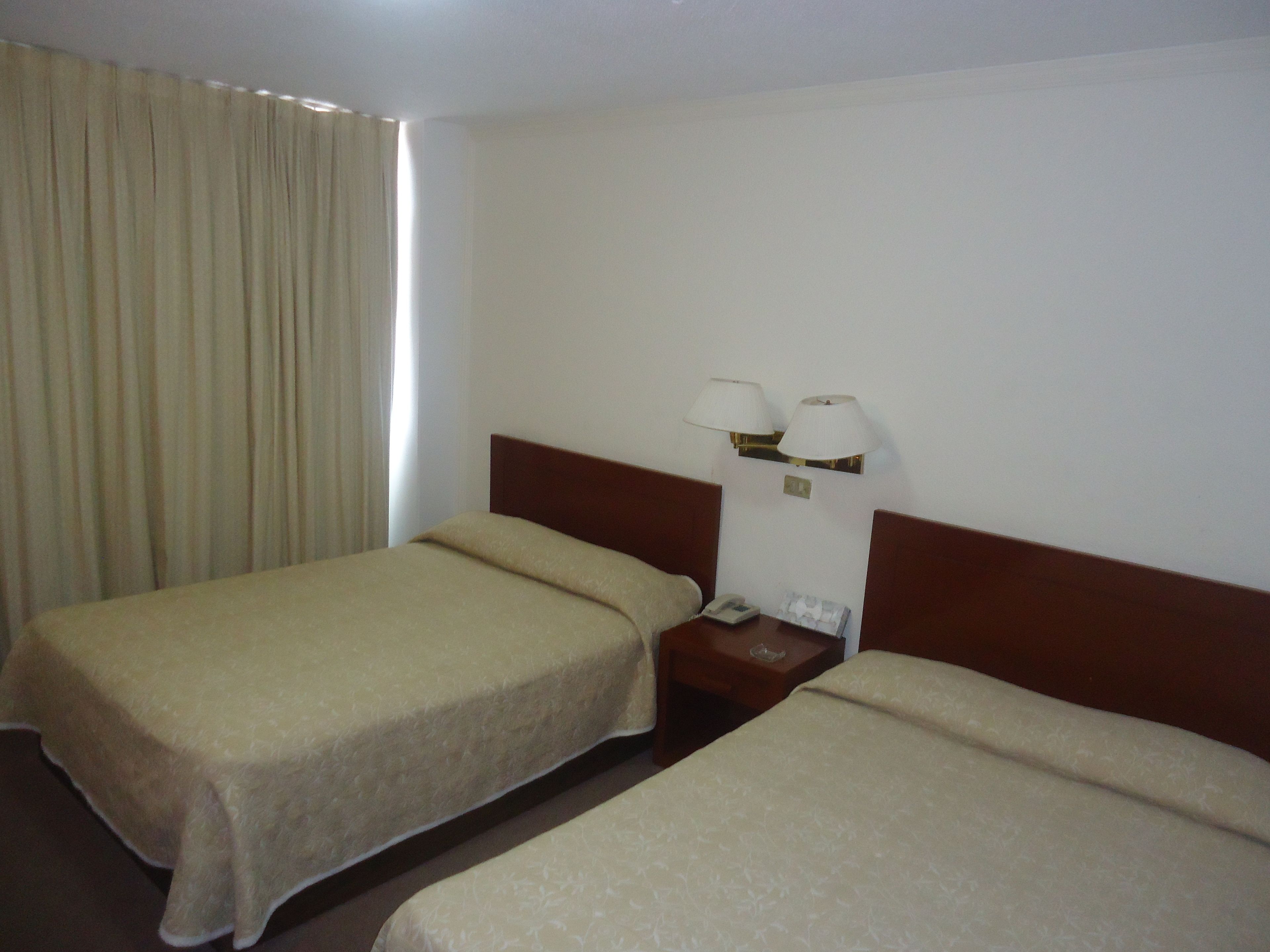 Una camera d'albergo con due letti singoli, un comodino con una lampada e una finestra con tende.