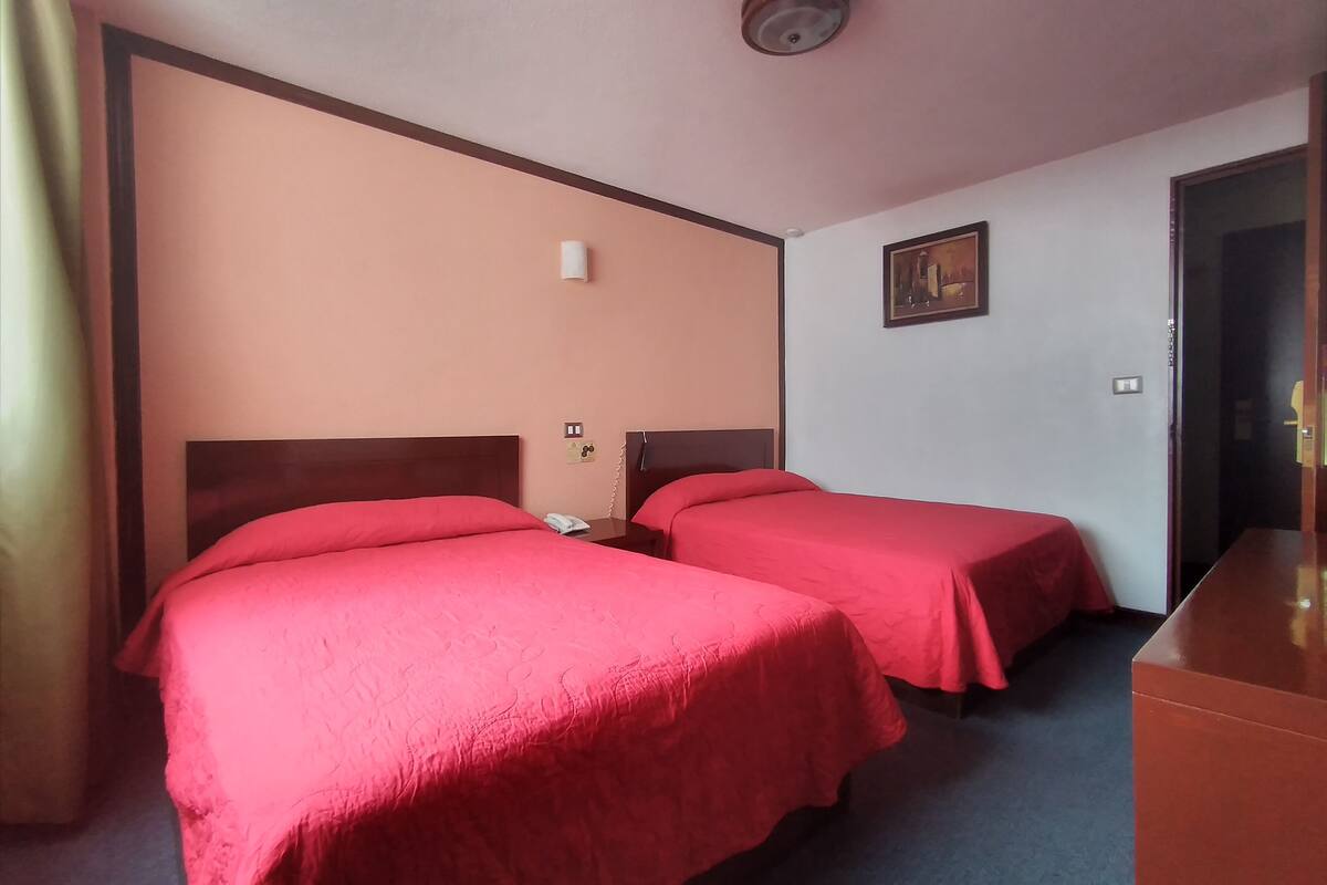A hotel room with two single beds, a desk, a chair, a telephone, and a painting on the wall.