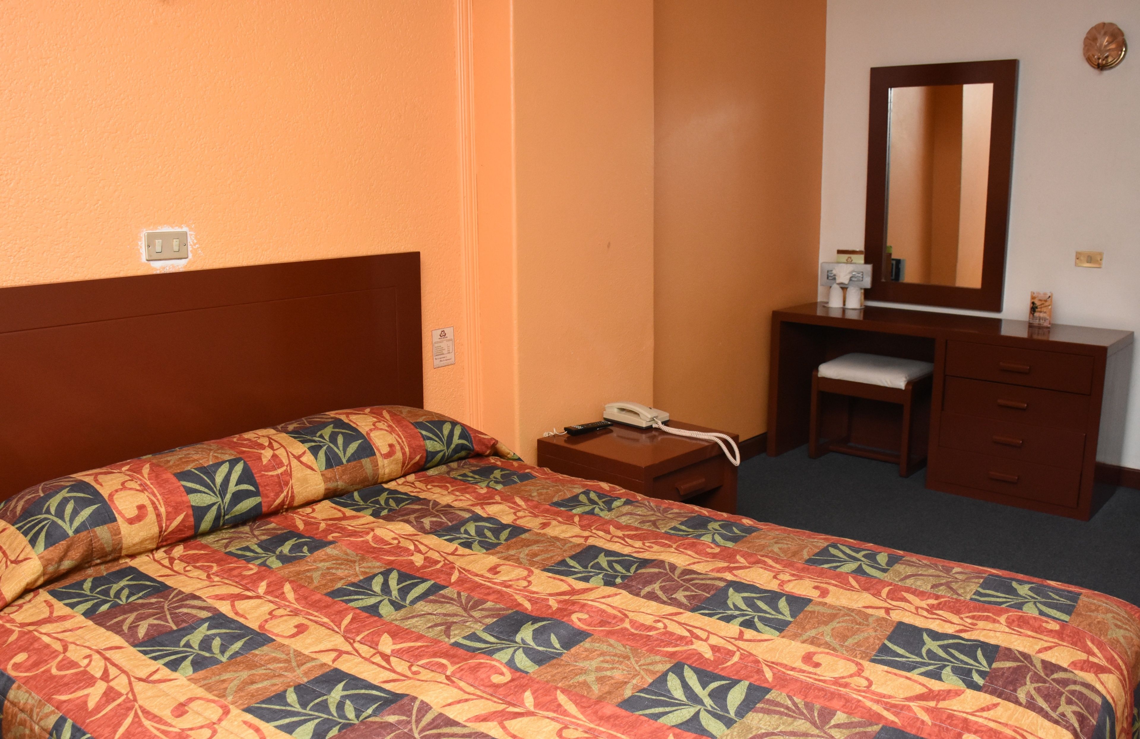 A hotel room with a large bed, a bedside table with a phone, a small desk, a mirror, and a dresser.