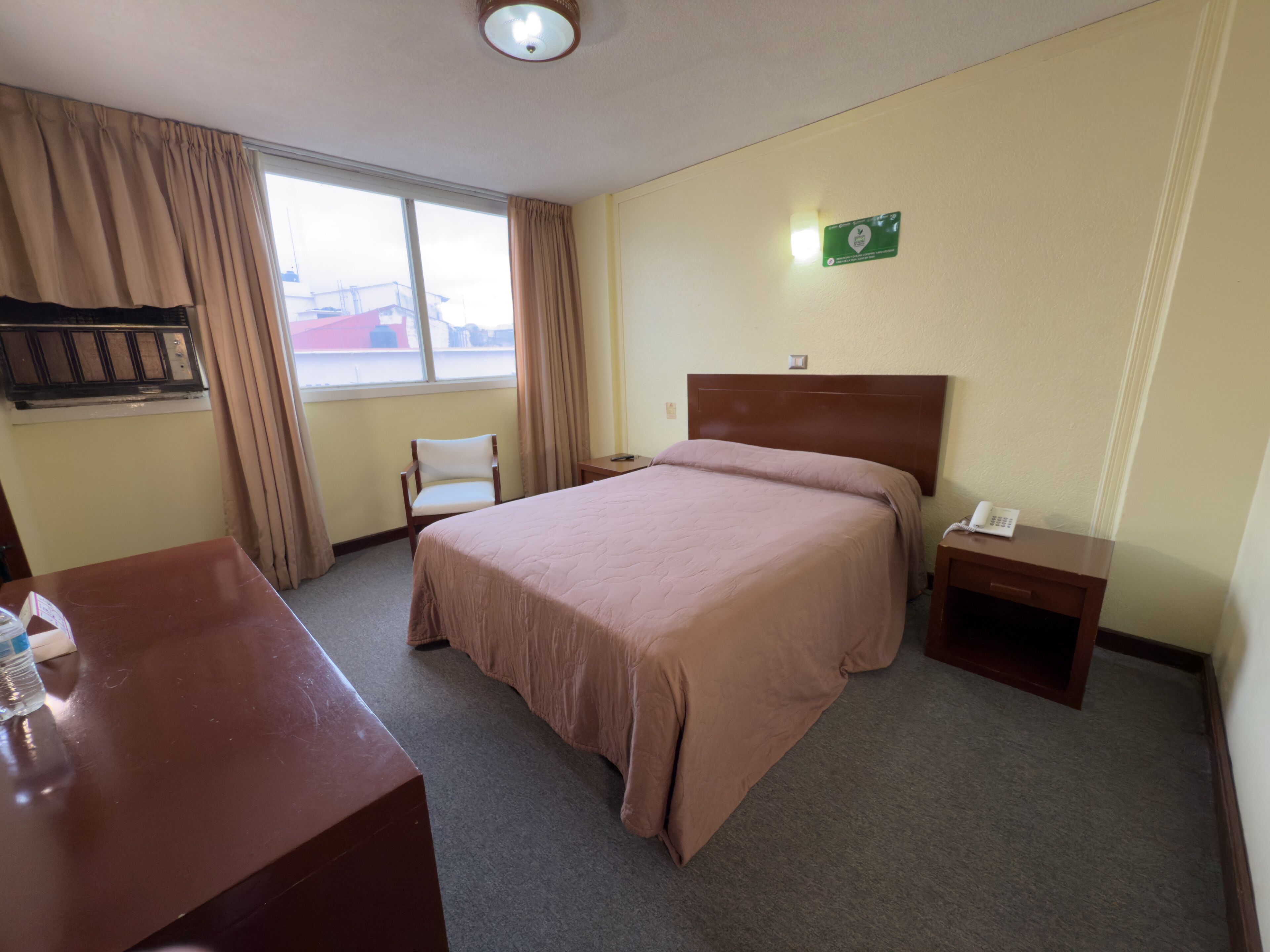 A hotel room with a large bed, a desk, a chair, a bedside table with a phone, and a window with curtains.