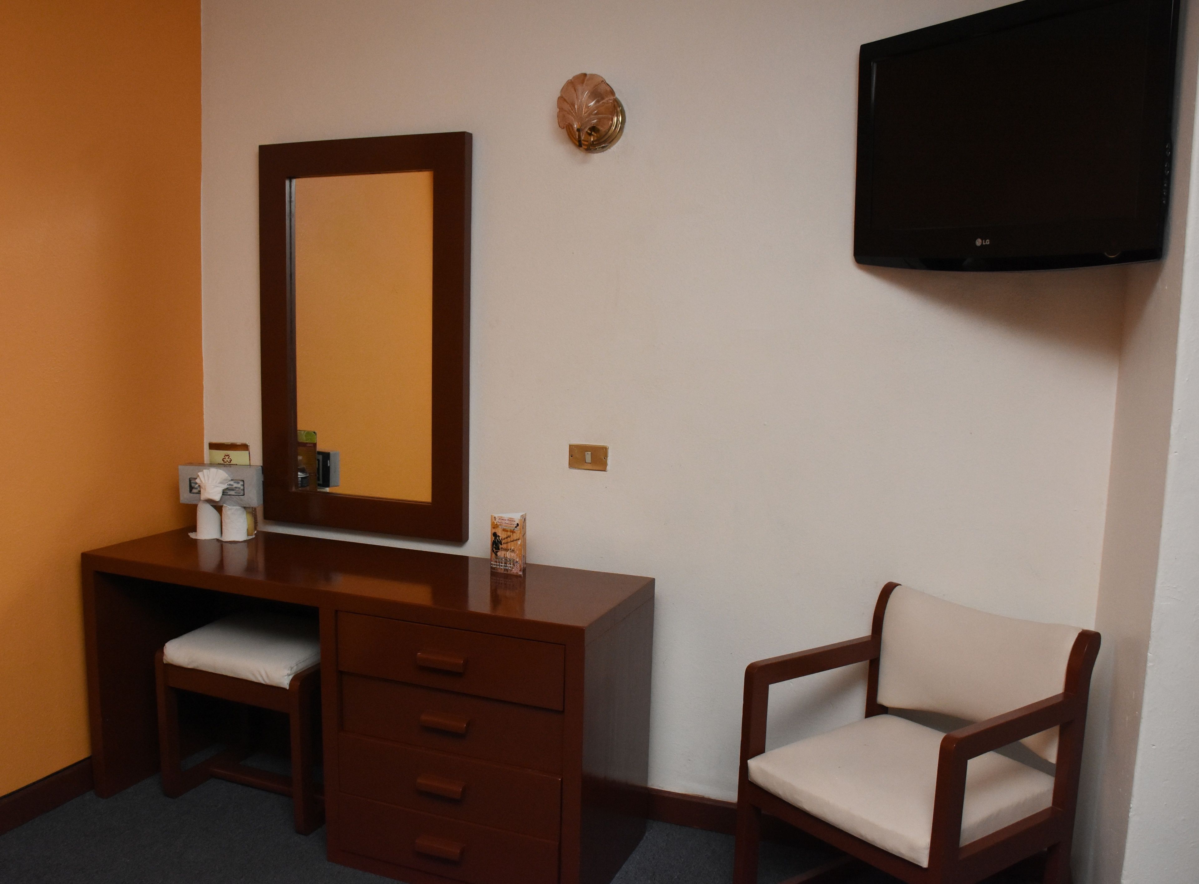 A hotel room with a wooden desk, a chair, a large mirror, and a flat-screen TV mounted on the wall.