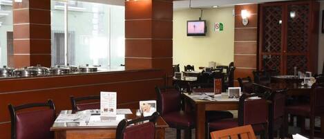 Un restaurante con mesas y sillas de madera, una zona de buffet con ollas, y un televisor en la pared.