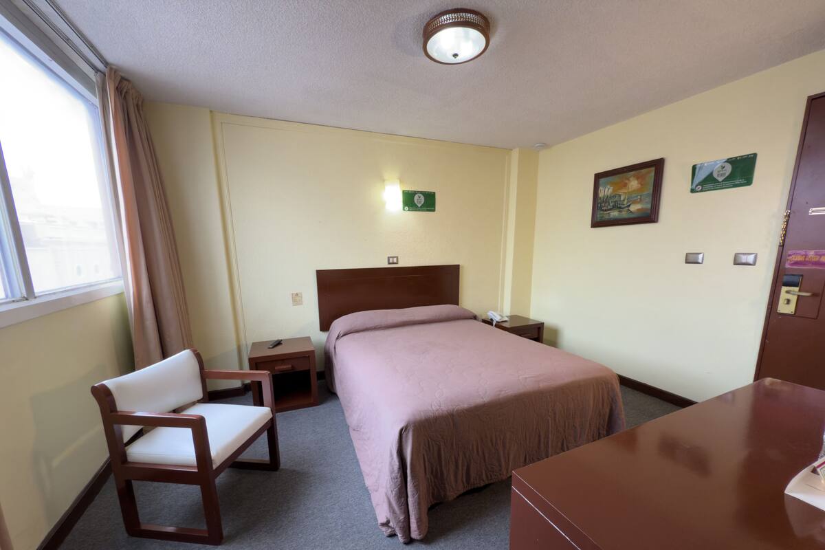 A hotel room with a bed, a desk, a chair, a nightstand, a lamp, and a window with curtains.