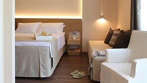 Habitación de hotel con cama, mesita de noche con lámpara, una silla y pantuflas en el suelo.