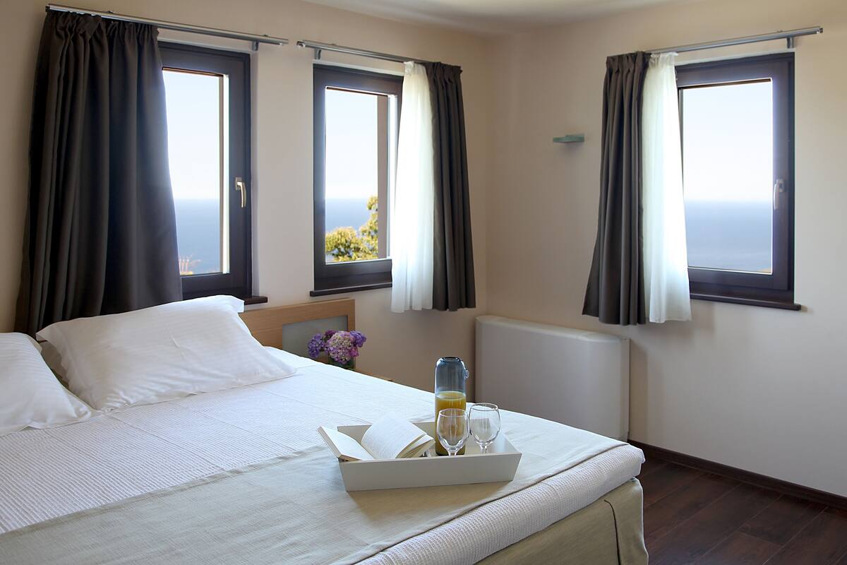 Quarto de hotel com cama, duas janelas, radiador e bandeja com sumo de laranja e dois copos.