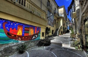 A narrow street with a colorful mural on the left wall, a staircase leading to an upper floor, and a cobblestone path.