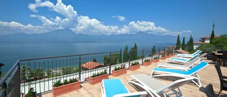 A terrace with sun loungers and potted plants, overlooking a body of water and mountains.