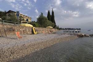 Una spiaggia di ciottoli con lettini e un kayak, un muro in pietra e un edificio con un balcone sullo sfondo.