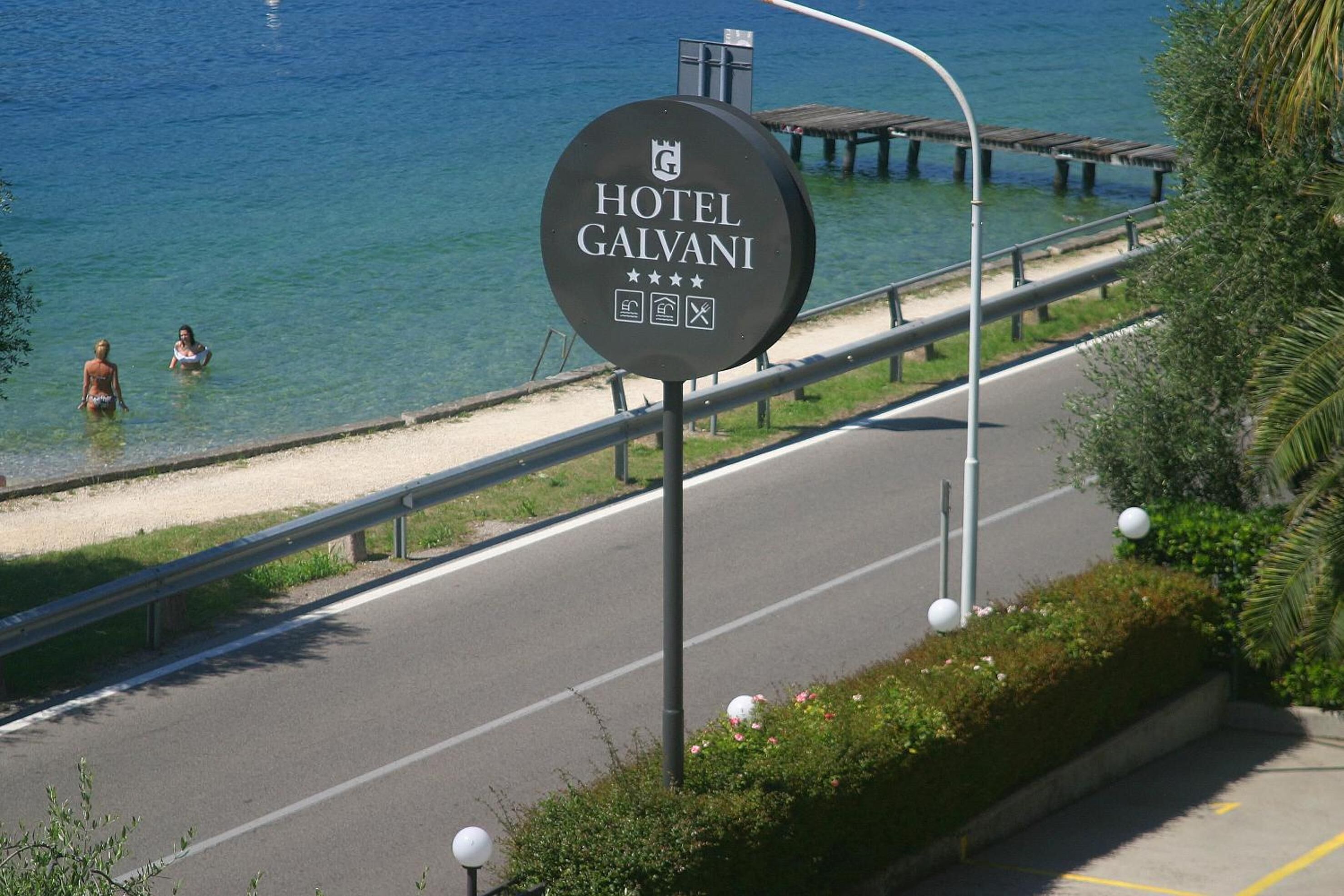 A sign for Hotel Galvani by the beach with clear blue water and a wooden pier.