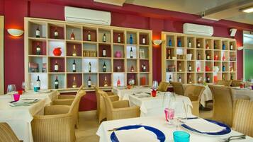 A restaurant with multiple tables, wicker chairs, and shelves of wine bottles.