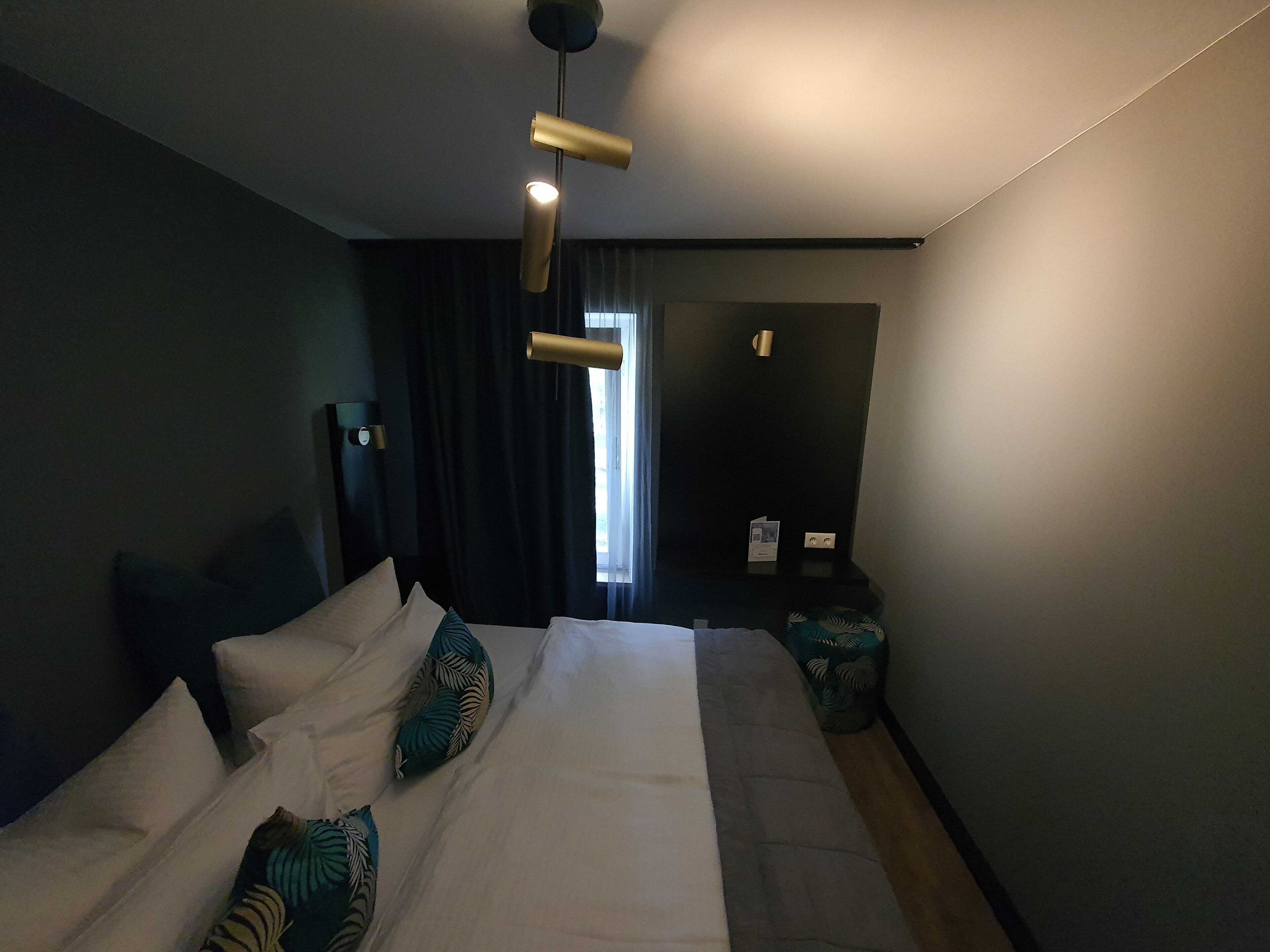 A modern hotel room with a large bed, a bedside table, a lamp, and a view of the outdoors.
