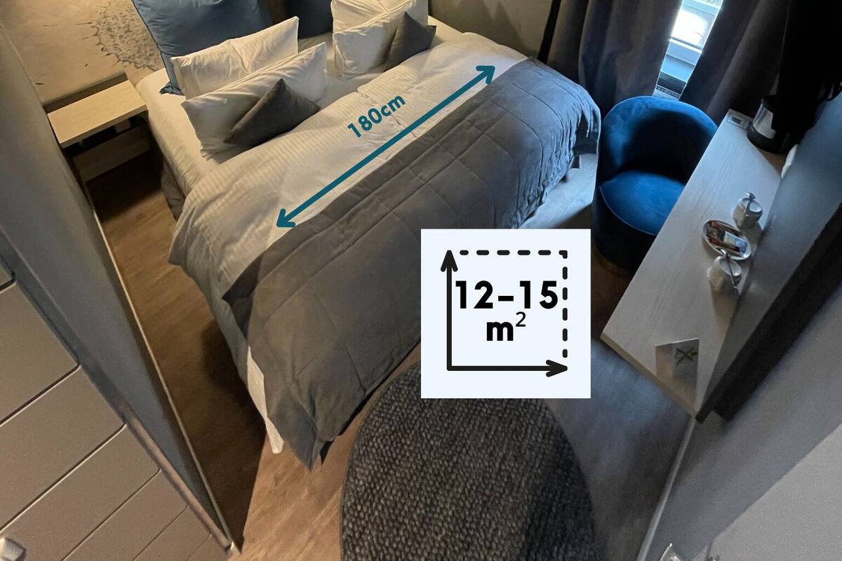 Habitació d'hotel senzilla amb un llit de 180 cm, una taula petita i una cadira. La habitació té un mural de peixos a la paret i una finestra amb cortines.