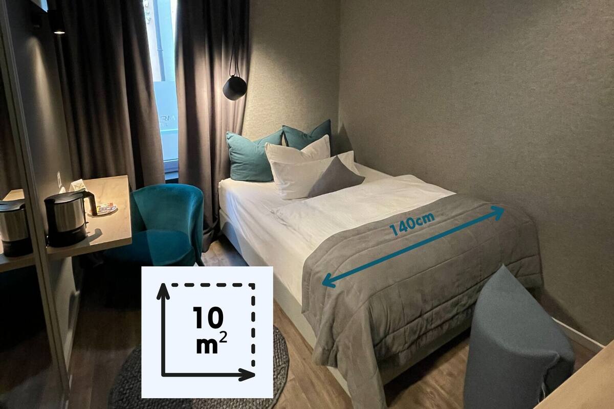 Habitació d'hotel amb un llit de 140cm, una taula petita amb una paperera, una cadira i una catifa rodona.