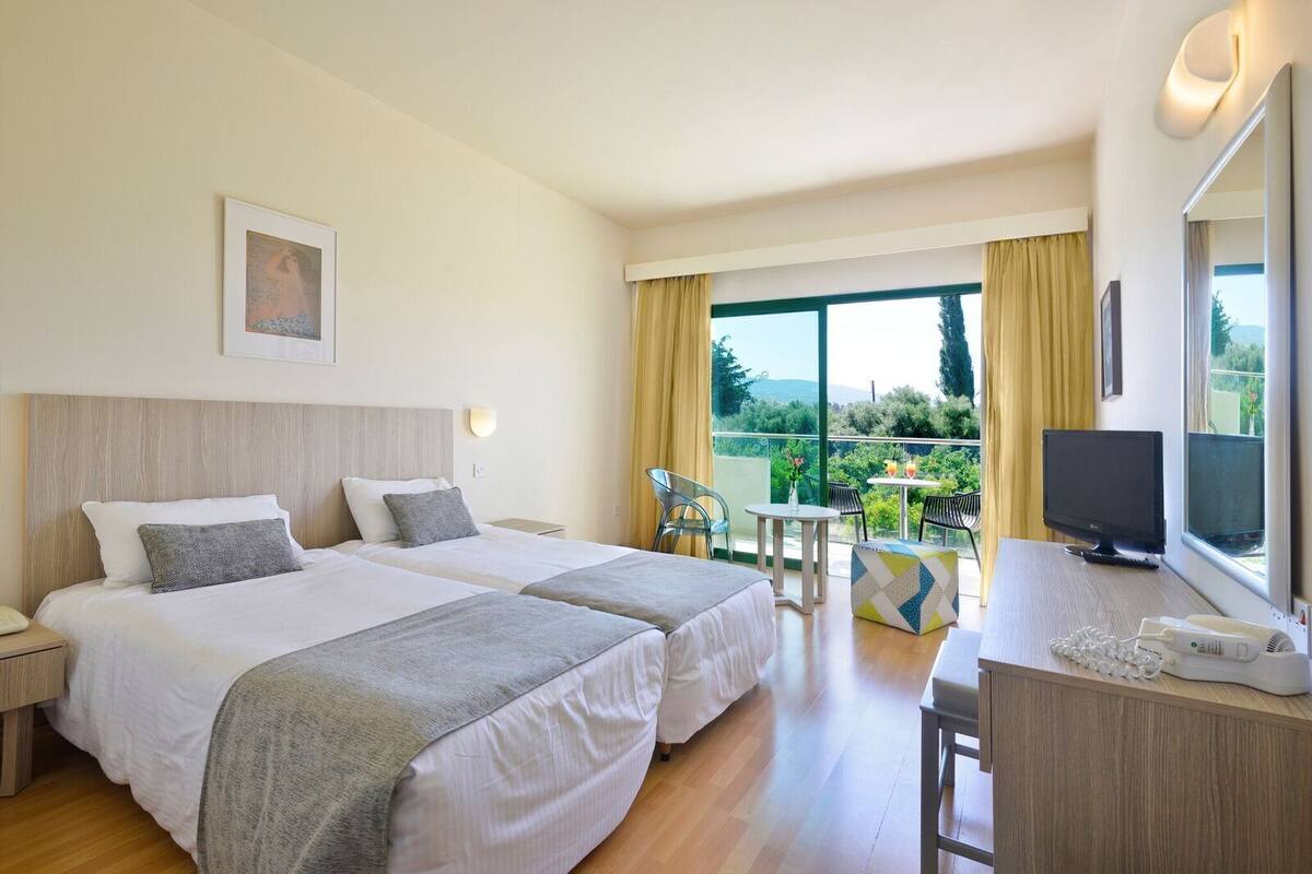 Une chambre d’hôtel avec deux lits, un balcon avec une table et des chaises, une télévision et un bureau avec un téléphone.