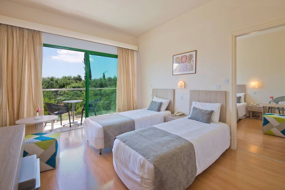 Une chambre d’hôtel avec deux lits, un balcon avec une table et des chaises, un petit bureau et un tableau au mur.