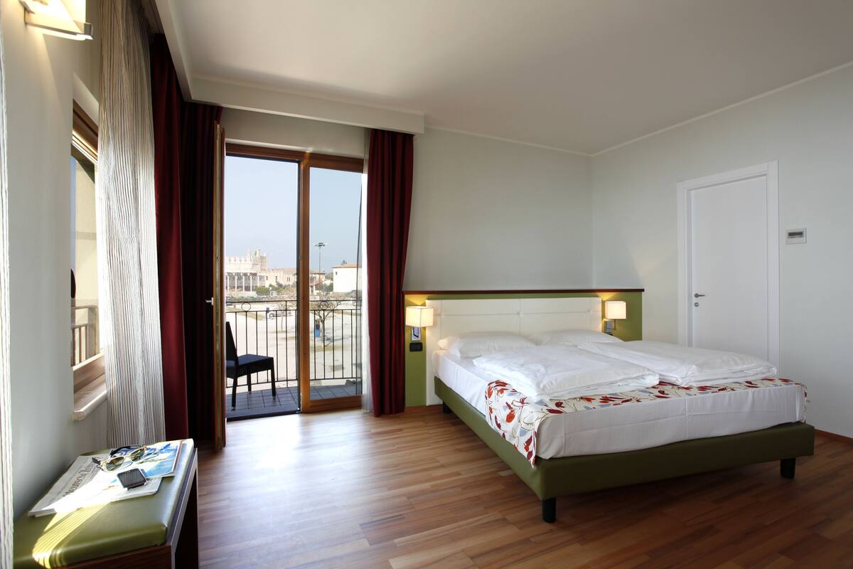 A hotel room with a bed, a desk with a chair, a window with curtains, and a view of a cityscape.