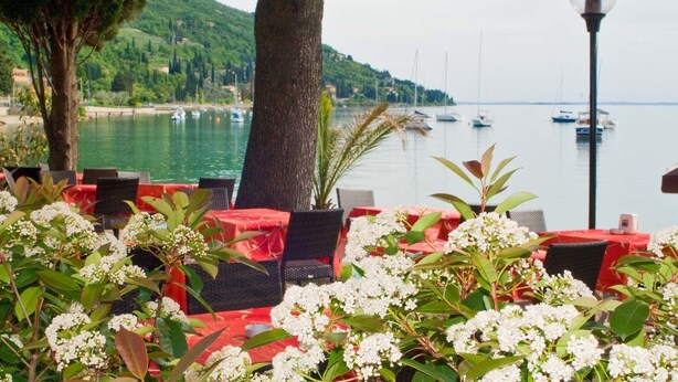 A terrace with red tables and chairs, overlooking a body of water with boats and greenery in the background. - Hotel Lido (Torri del Benaco)