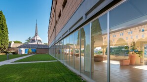 A modern building with a glass facade, a grassy courtyard, and a church with a tall steeple in the background. - Villa EUR - Parco dei Pini (Rome)