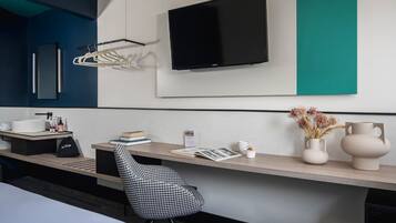 Une chambre d’hôtel moderne dotée d’un grand téléviseur à écran plat fixé au mur, d’un bureau en bois avec une chaise, d’un vase blanc contenant des fleurs séchées et d’un magazine.