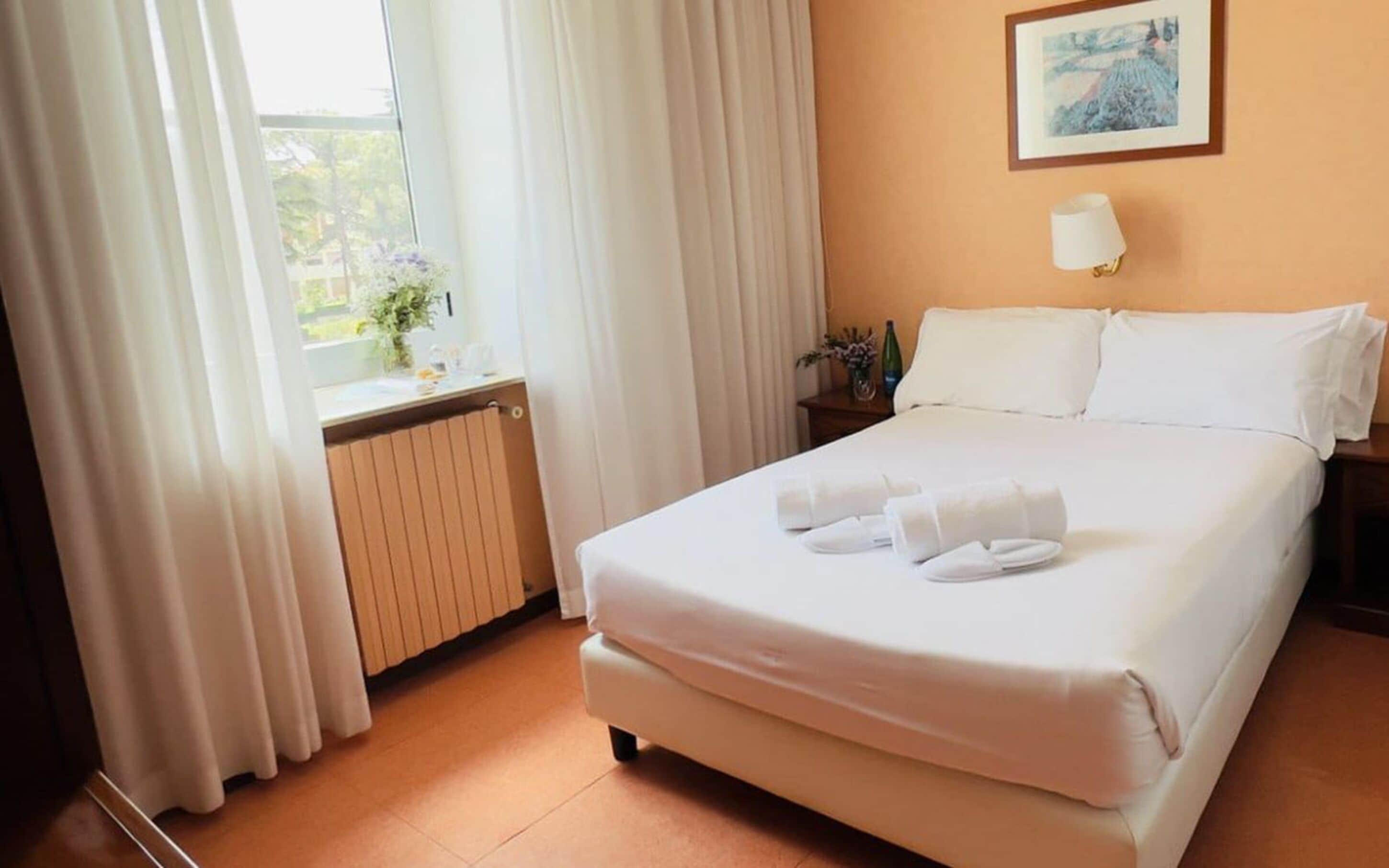 A hotel room with a bed, white curtains, a radiator, a nightstand with a lamp, and a framed picture on the wall.