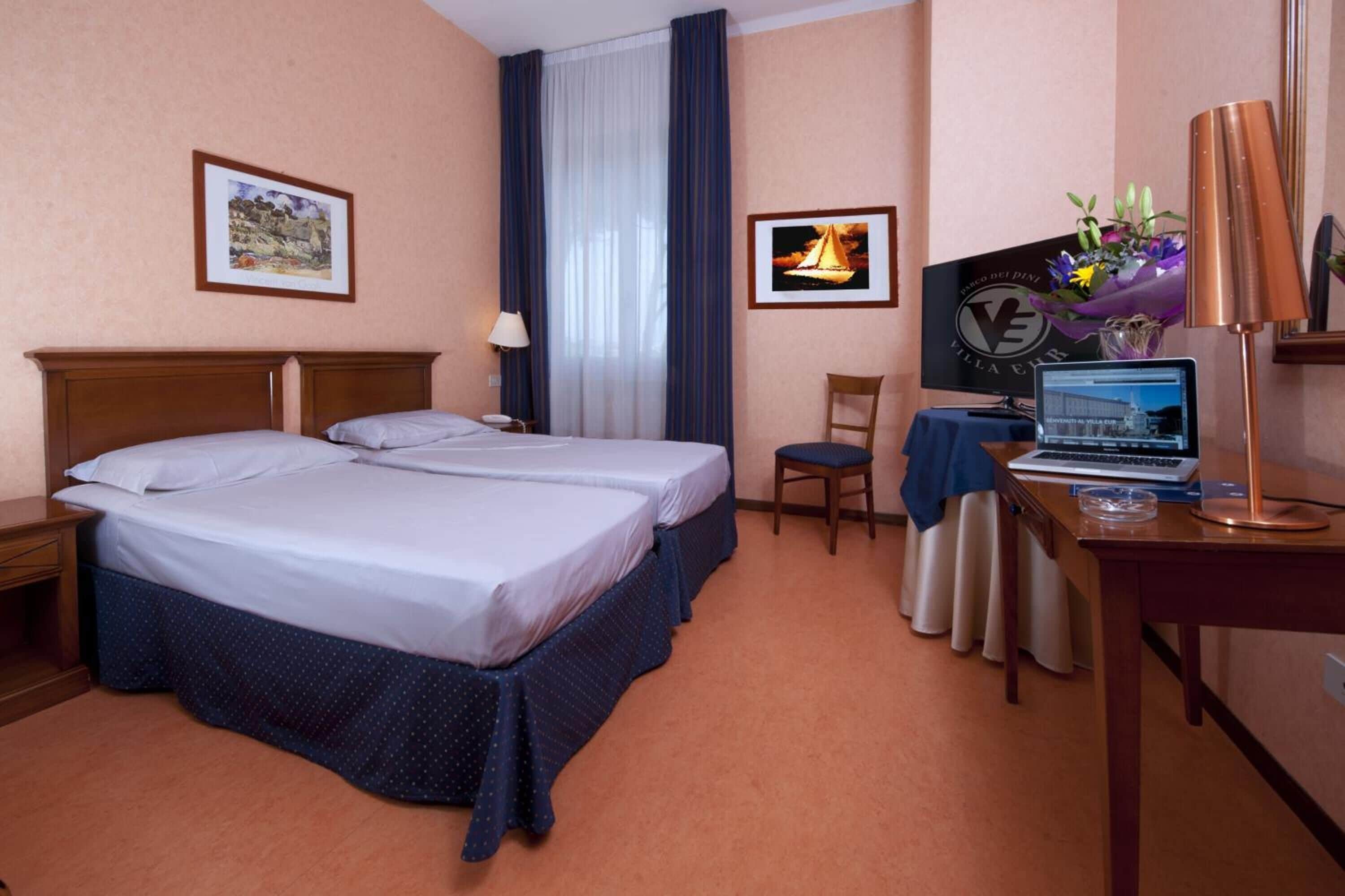 Camera d'albergo con due letti, una scrivania con un portatile, una sedia, una lampada e quadri alle pareti.