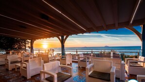 Eine Terrasse mit weißen Korbmöbeln und einem gemusterten Boden, mit Blick auf einen Sonnenuntergang über dem Meer.