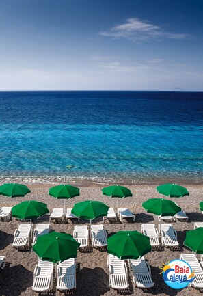 A beach with rows of white lounge chairs and green umbrellas, a clear blue sea, and a logo in the bottom right corner. - Villaggio Baia Calavà Hotel e Residence (Gioiosa Marea)