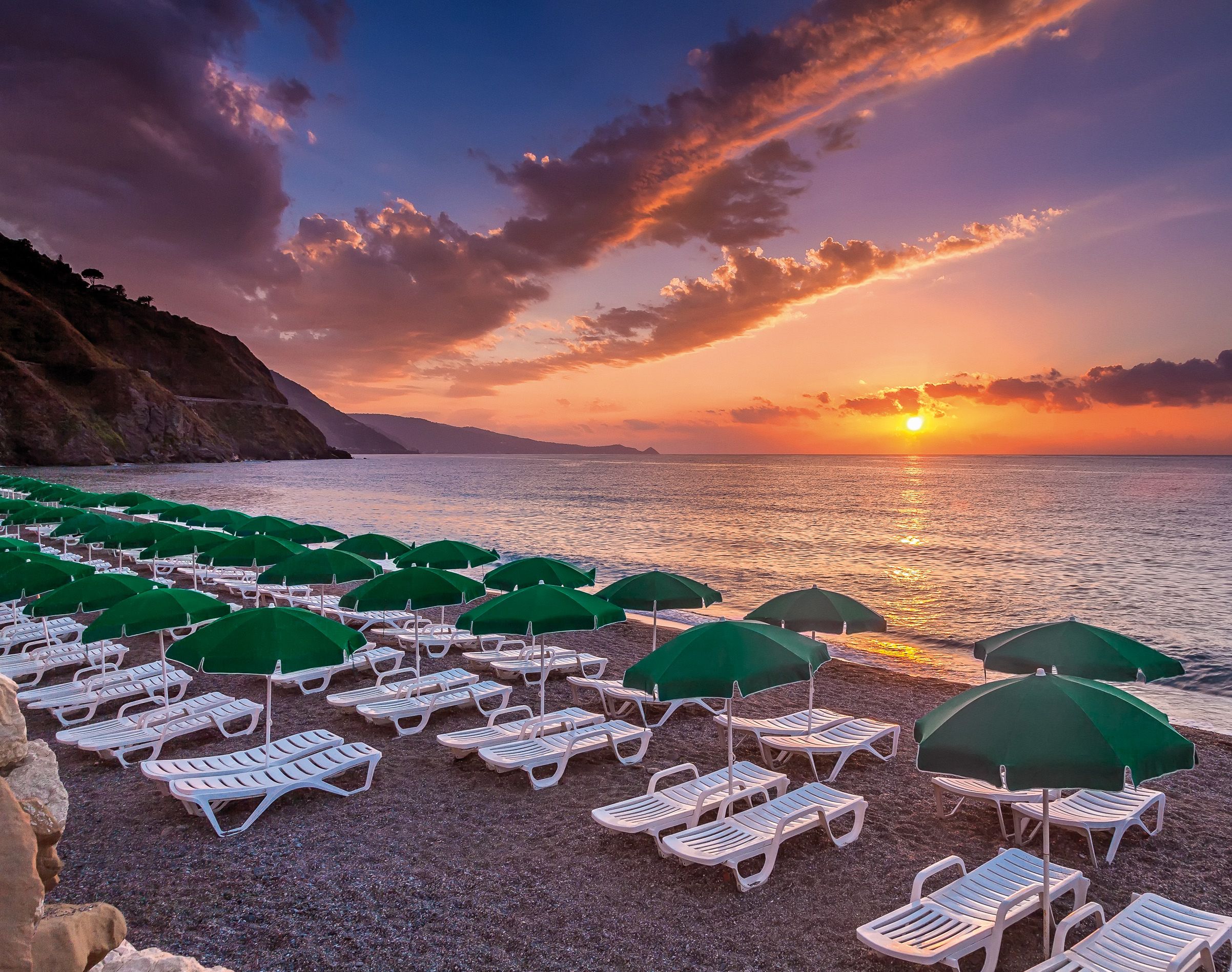 Una spiaggia con file di sdraio bianche e ombrelloni verdi, un tramonto sull'oceano e scogliere rocciose sullo sfondo.
