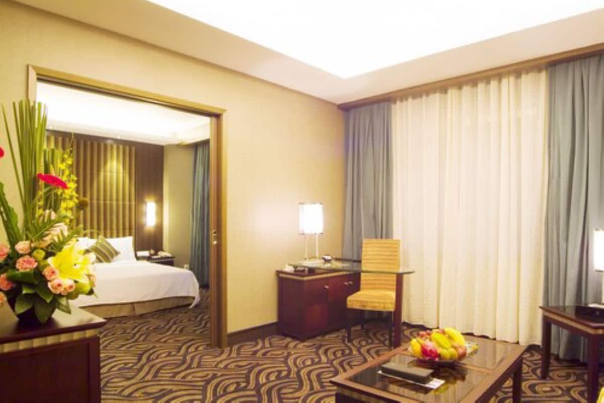 Habitación de hotel con cama, escritorio con lámpara, una silla, una mesita de centro con fruta y un ventanal con cortinas.