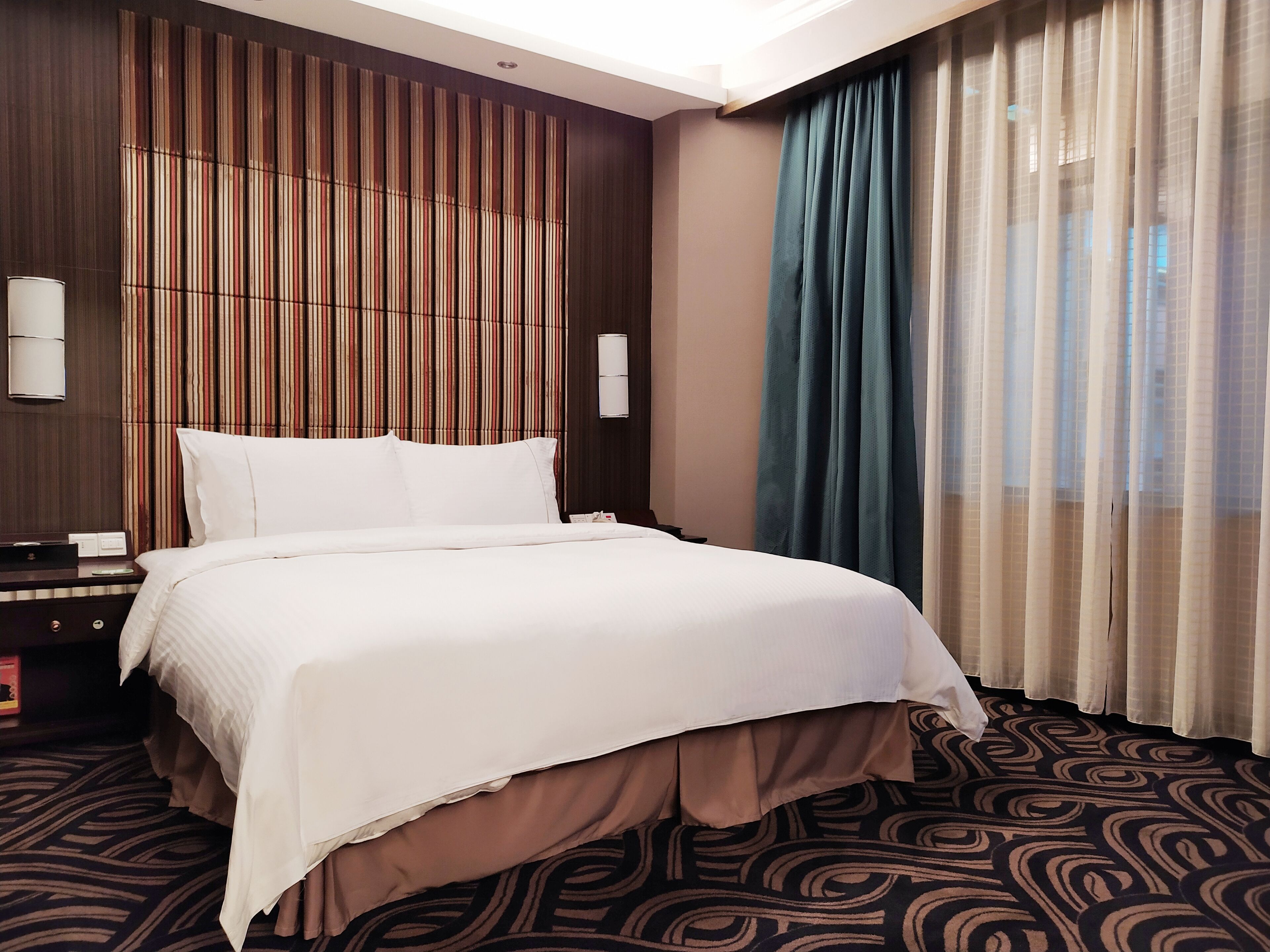 Habitación de hotel con una cama grande, una mesita de noche, una lámpara, luz empotrada en la pared y una ventana con cortinas.