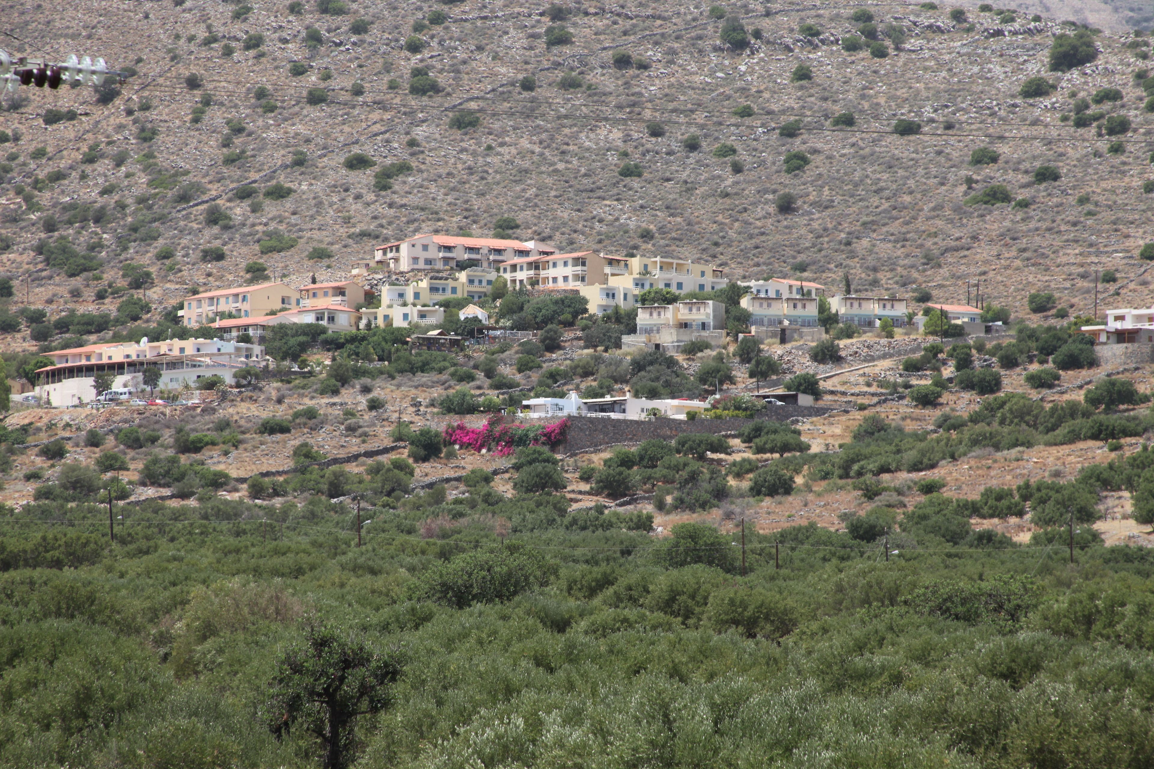 A hillside residential area with multiple buildings, some with red roofs, surrounded by sparse vegetation and rocky terrain.