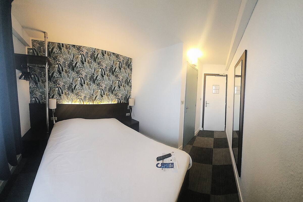 A hotel room with a bed, a headboard with a leaf pattern, a nightstand, a lamp, a mirror, and a door leading to another room.