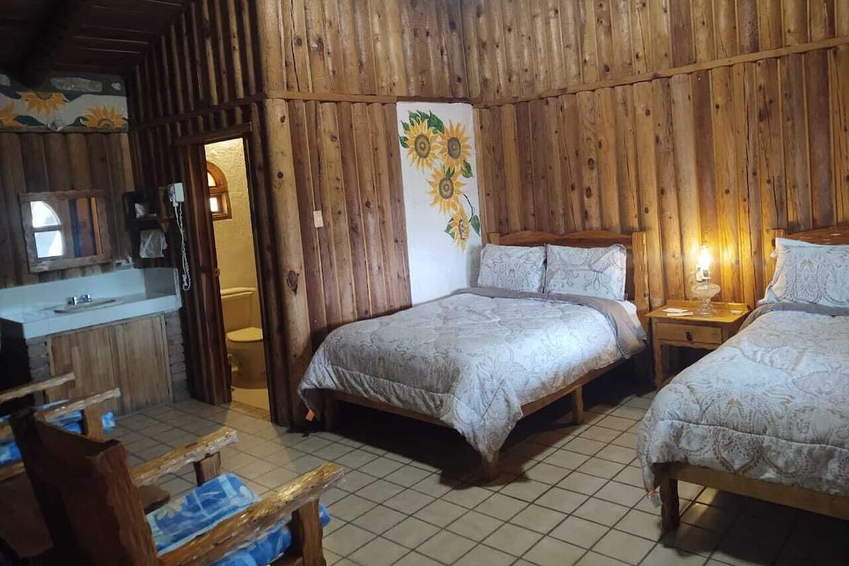 A rustic wooden room with two beds, a small table, and a bathroom visible through an open door.