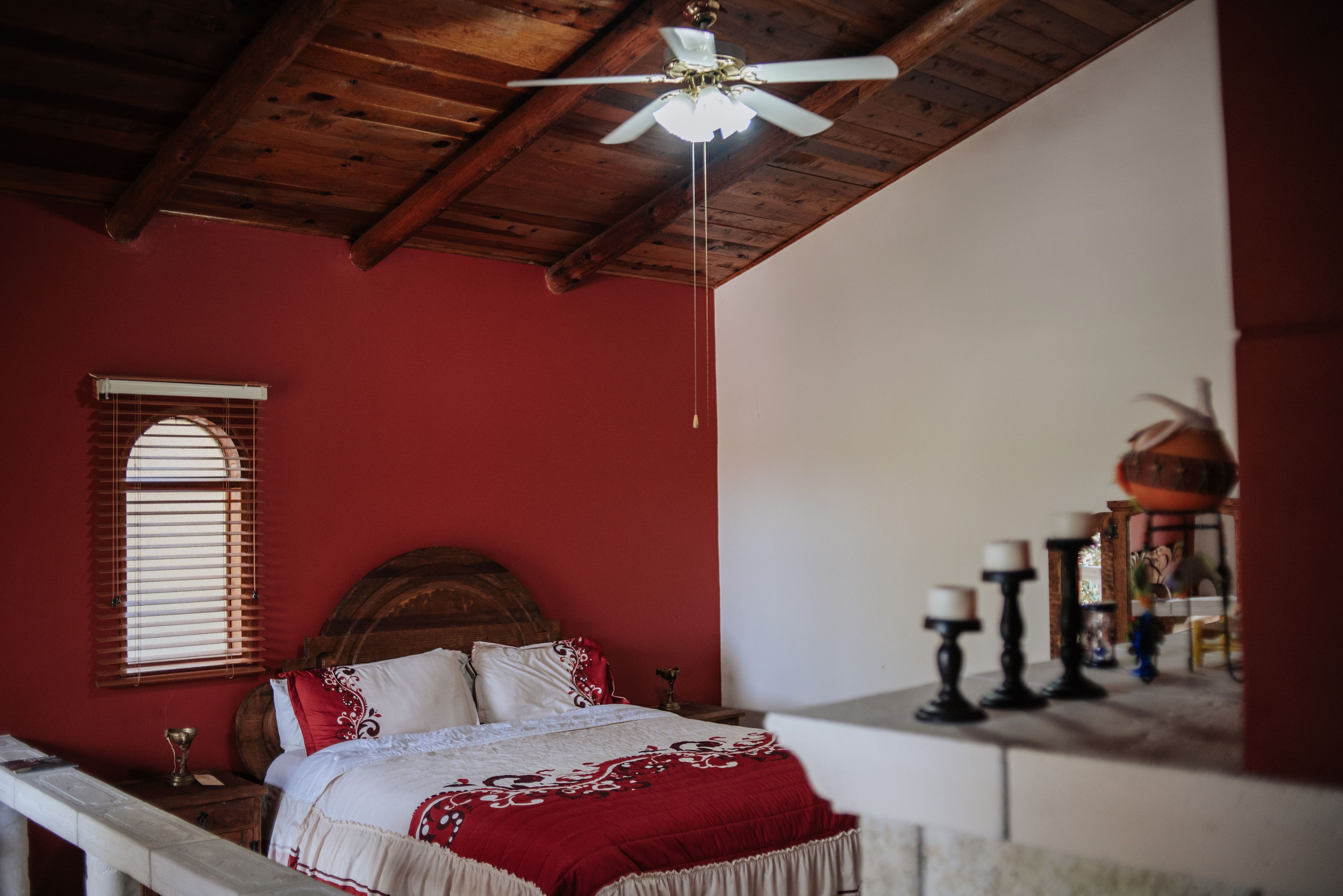 A bedroom with a wooden ceiling, a bed with white and red bedding, a window with blinds, and a nightstand with a lamp.