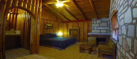 A cozy log cabin room with a bed, a small table, a chair, a stone wall, and a fireplace.