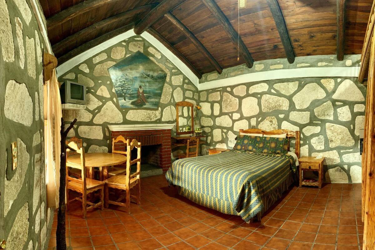 A rustic bedroom with a stone wall, wooden ceiling beams, a bed, a wooden chair, a small table, and a fireplace.